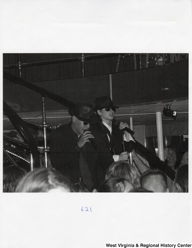 Two unidentified young men sing into microphones at a party for a crowd.