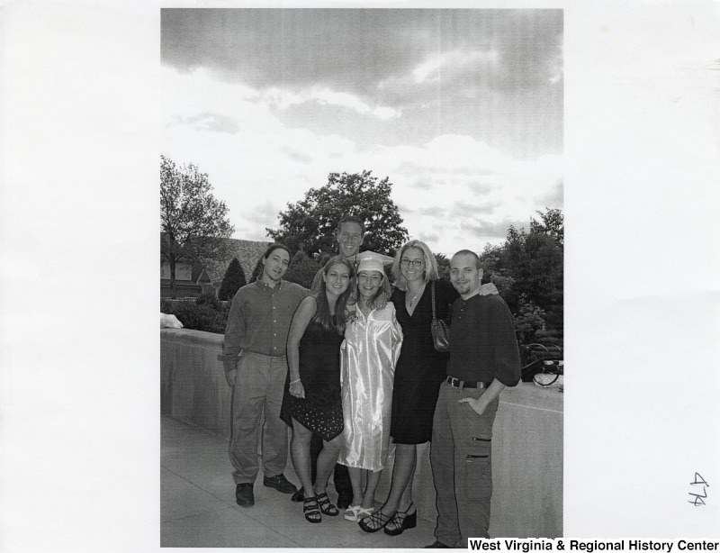 Three unidentified men and three unidentified women stand together for a photograph outside. One young woman wears a graduation gown and cap.
