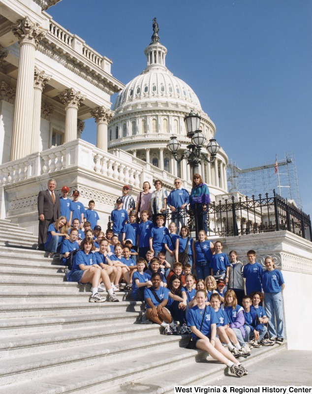 On the far left, Representative Nick J. Rahall (D-W.Va.) stands with an unidentified group of children all wearing blue shirts in front of the Capitol building.
