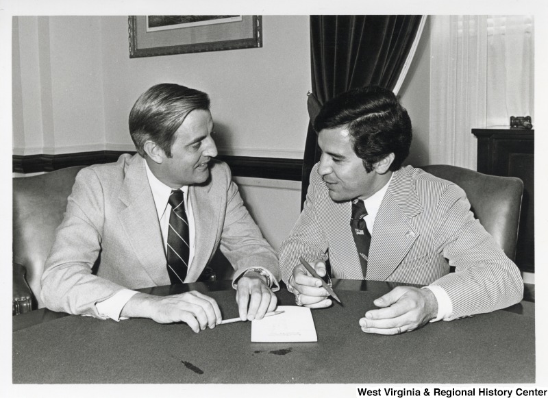 On the right, Representative Nick J. Rahall (D-W.Va.) sits and talks with Vice President Walter Mondale (D-MN) as they hold pens near paper.