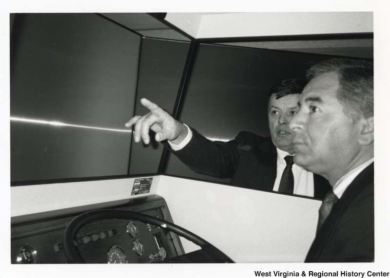 Representative Nick J. Rahall (D-W.Va.) sits behind the wheel of a vehicle while an unidentified man stands outside and points.