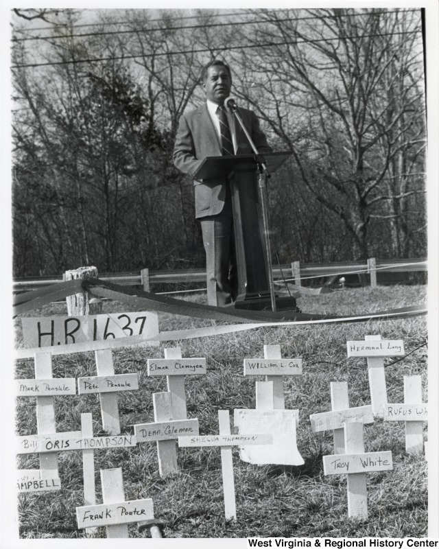 Representative Nick J. Rahall (D-W.Va.) stands behind a podium outside and gives a speech behind rows of white crosses with names on them for a Black Lung Ceremony.